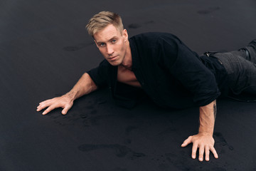 Wall Mural - Portrait of handsome blue-eyed man in black shirt lying on black sand beach