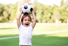 Excited Little Toddler Boy Playing Football On Soccer Field Outdoors: The Kid Is Holding Ball Above Head And Shouting Ready To Throw It. Active Childhood And Sports Passion Concept