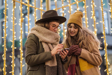 Young Women Using A Phone On The Street With Christmas Decoration Behind Them
