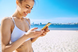 Fototapeta  - Closeup portrait of beautiful sexy happy girl on the sand city beach with sea background. Young smiling woman standing with the smartphone in the hand, looking at phone against blue sky and sea