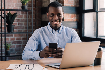 Wall Mural - African man spending time in office with phone in hand, smiling at camera and feeling confident