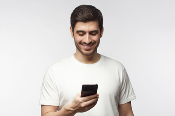 Wall Mural - Closeup of Caucasian man holding phone in one hand and smiling happily while looking at screen, isolated on grey background