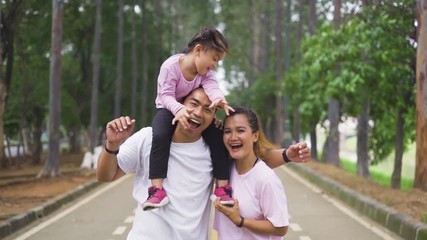 Wall Mural - Slow motion of cheerful family smiling on the camera with father giving daughter piggyback ride on his shoulders at the park. Shot outdoors