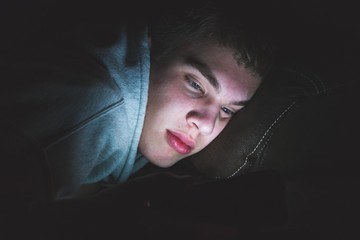 Wall Mural - Teenager lying down on a couch in the dark. The light from the screen of his smartphone is illuminating his face.