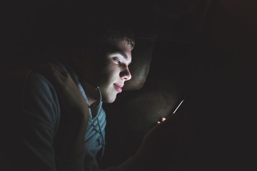 Wall Mural - Teenager lying down on a couch in the dark. The light from the screen of his smartphone is illuminating his face.