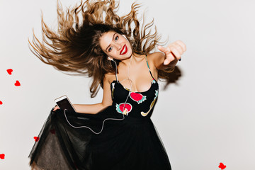 Winsome long-haired girl dancing on light background and expressing happiness. Graceful brunette lady in dress having fun in studio decorated with hearts.