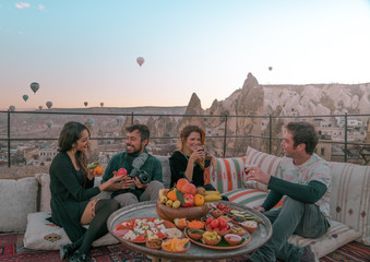 Four friends or two couples have fun whilst having breakfast outdoor with fruits, breads and drinks with a beautiful view to the balloons in Cappadocia, Turkey 