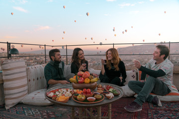 Four friends or two couples have fun whilst having breakfast outdoor with fruits, breads and drinks with a beautiful view to the balloons in Cappadocia, Turkey 