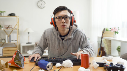 Wall Mural - Man addicted to computer games. concentrated young asian male in headset sitting in dirty apartment having fun on internet using keyboard staring pc screen monitor. technology cause eyes near sight