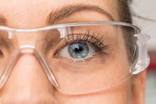 A Beautiful Young Caucasian Is Viewed Closeup In The Workplace, Wearing Protective Goggles Over Her Eyes. Pretty Eyelashes And Blue Eyelashes Seen In Detail Behind Plastic Eyewear.