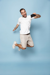 Wall Mural - Full length photo of unshaved caucasian man in casual white t-shirt jumping and taking selfie on smartphone