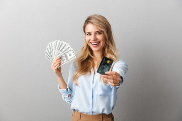 Wall Mural - Happy young pretty blonde business woman posing isolated grey wall background holding credit card and money.