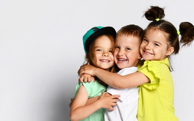 Studio portrait of children on a light background: full body shot of three children in bright clothes, two girls and one boy. Triplets, brother and sisters. hugging on camera. Family ties, friendship