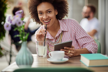 Wall Mural - Gorgeous mixed race woman in pink striped dress drinking lemonade and using tablet while sitting in cafe.