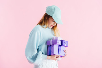 Wall Mural - excited pretty teenage girl in cap looking at purple gifts isolated on pink