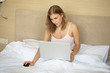© lashkhidzetim - Smiling woman catching up on her social media as she relaxes in bed with a laptop computer on a lazy day.