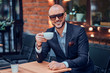 © Fxquadro - Elegant smiling man is drinking coffee while sitting outside at cafe on his break.