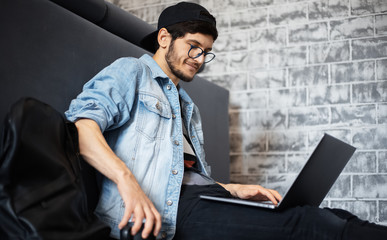 young man in denim jacket and black cap sitting on the floor working on laptop