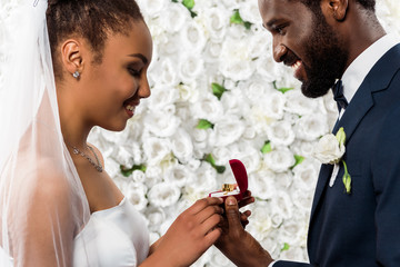 cheerful african american bride looking at box with ring near happy bridegroom and flowers