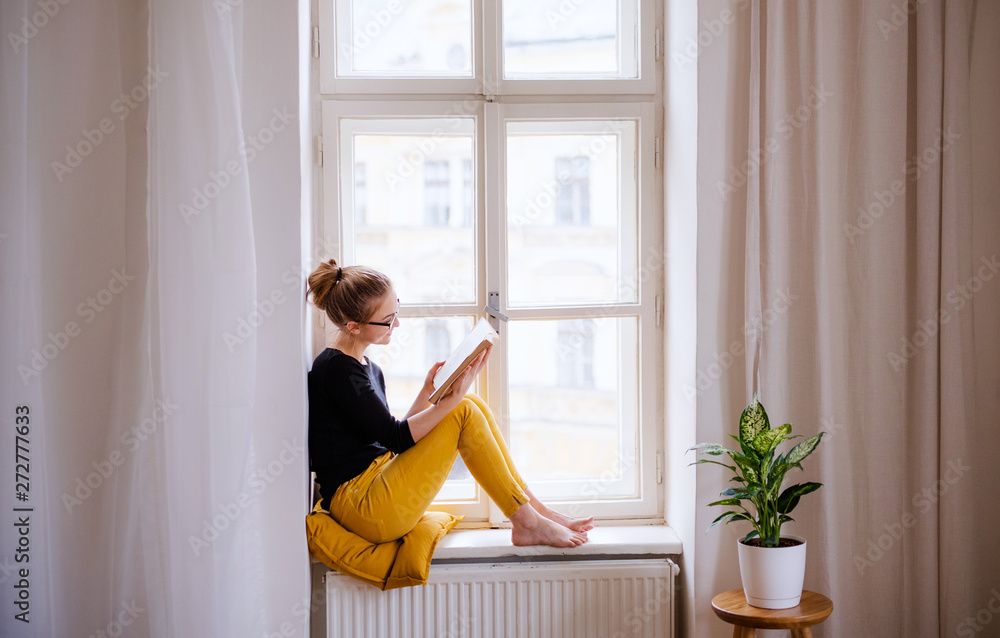 A young female student with a book sitting on window sill, studying. - obrazy, fototapety, plakaty 