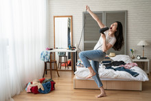 Full Length Of Funny Asian Girl Dancing And Singing With Hair Dryer In Wood Cozy Bedroom At Home. Young Chinese Woman Crazy Joyful With Clothes Dresses On Bed Prepared For Luggage On Sunshine Day.