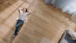 Young Woman is Lying on a Wooden Flooring in an Apartment. She's Dressed Casually. Cozy Living Room with Modern Minimalistic Interior and Wooden Parquet. Top View Camera Shot.