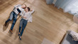 © Gorodenkoff - Young Couple are Lying on a Wooden Flooring in an Apartment. They are Using a Tablet Computer. Cozy Living Room with Modern Interior, Grey Sofa and Wooden Parquet. Top View Camera Shot.