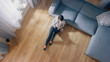 © Gorodenkoff - Young Woman is Sitting on a Floor, Working or Studying on a Laptop. Cozy Living Room with Modern Interior, Grey Sofa and Wooden Flooring. Top View Camera Shot.