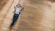 © Gorodenkoff - Young Man is Lying on a Wooden Flooring in an Apartment. He's Wearing a Jacket and White Shirt. Cozy Living Room with Modern Minimalistic Interior and Wooden Parquet. Top View Camera Shot.