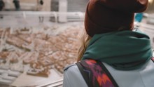 Young Female Tourist With A Backpack On Her Shoulders Examines A Miniature Installation Of The City With Houses And Streets. Slow Motion.