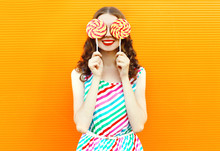 Portrait Happy Smiling Woman Hiding Her Eyes With Two Lollipop In Colorful Striped Dress On Orange Wall Background