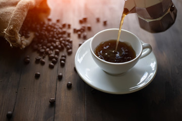 that hand is Pouring coffee from the Italian coffee maker(mocha pot) into a white ceramic coffee cup Placed on a brown wood table there are roasted coffee beans, sag in the morning