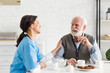 © LIGHTFIELD STUDIOS - Nurse sitting on kitchen with senior man, putting hand on his shoulder