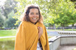 © Mangostar - Smiling woman wrapping in coat in park. Young lady looking at camera with blurred green tree and river in background. Nature and leisure concept. Front view.