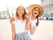 Portrait of two young beautiful blond smiling hipster girls in trendy summer white t-shirt clothes. Sexy carefree women posing on street background. Positive models having fun in sunglasses and hat