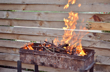 Dry wood for grilling burn with a large flame on the grill, against a wooden fence