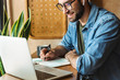 © Drobot Dean - Photo of masculine bearded man writing notes and using laptop while working in cafe indoors