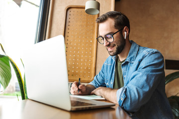 Poster - Photo of handsome bearded man writing notes and using laptop while working in cafe indoors