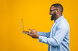 © denis_vermenko - Young smiling african american man standing and using laptop computer isolated over yellow background.