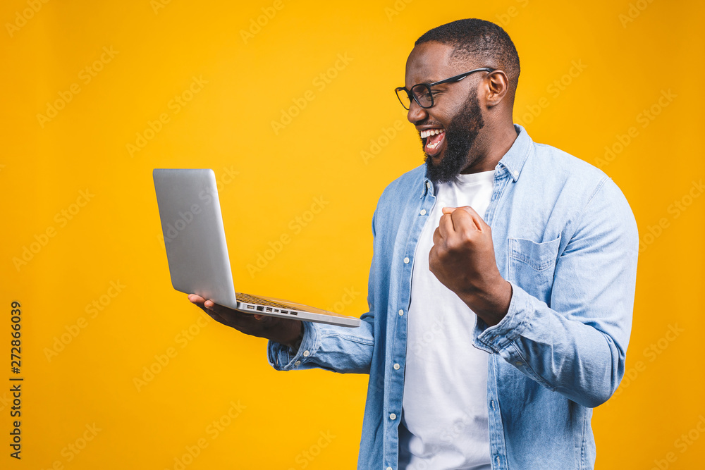 Excited happy afro american man looking at laptop computer screen and celebrating the win isolated over yellow background.