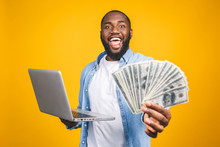 Photo Of A Happy Young Afro American Handsome Man Posing Isolated Over Yellow Wall Background Using Laptop Computer Holding Money.