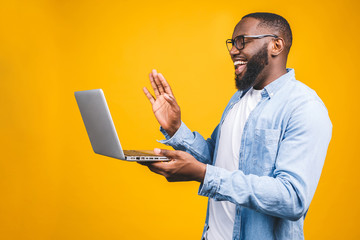 Young happy smiling african american man standing and using laptop computer isolated over yellow background.
