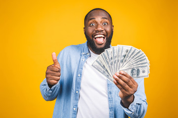 Winner! Young rich african american man in casual t-shirt holding money dollar bills with surprise isolated over yellow wall. Thumbs up.