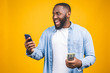 © denis_vermenko - Winner! Young rich african american man in casual t-shirt holding money  dollar bills and mobile phone with surprise isolated over yellow wall.