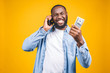© denis_vermenko - Winner! Young rich african american man in casual t-shirt holding money  dollar bills and mobile phone with surprise isolated over yellow wall.