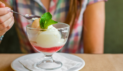 young girl's hand holding a spoon and  eating ice cream in glass at a table in cafe