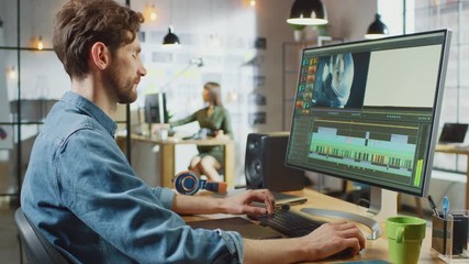 Wall Mural - male video editor with beard and jeans shirt works with footage on his personal computer with big di