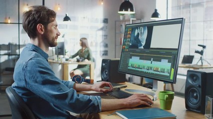 Wall Mural - male video editor with beard and jeans shirt works with footage on his personal computer with big di