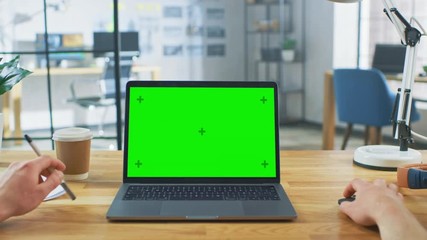 Canvas Print - Point of View Shot: Man Uses Laptop Computer with Green Mock-up Screen while Sitting at His Desk. He Holds Mouse and Pen in His Hands. In the Background Stylish Modern Open Space Office