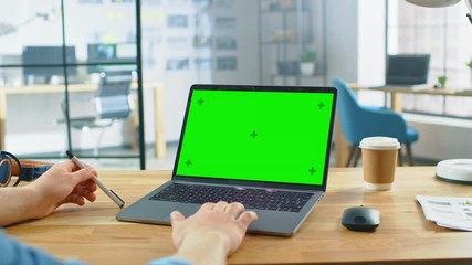 Point of View Shot: Man Uses Laptop Computer with Green Mock-up Screen while Sitting at His Desk. He Holds Mouse and Pen in His Hands. In the Background Stylish Modern Open Space Office